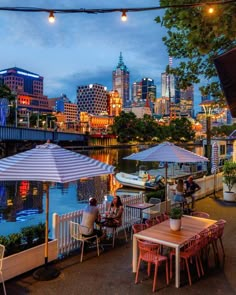 an outdoor dining area with tables and umbrellas overlooking the water at night, in front of a cityscape