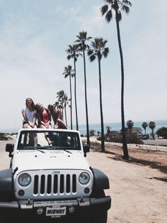 three people sitting on top of a white jeep in front of palm trees and the ocean