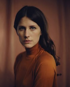 a woman in an orange turtle neck sweater looking at the camera with serious expression on her face