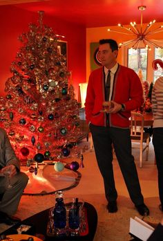 two men standing next to a christmas tree in a room with red walls and lights