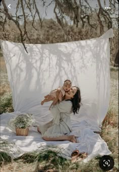 a woman holding a baby in her arms while sitting on a blanket under a tree