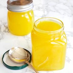 two jars filled with yellow liquid next to a spoon on a marble counter top,