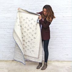 a woman holding up a white quilt in front of a brick wall with her hands on it