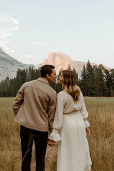 a man and woman holding hands walking through tall grass with mountains in the background at sunset