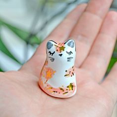 a small cat figurine sitting on someone's hand in front of a plant