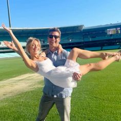 a man holding a woman on his back in front of a baseball field with an empty bleachers