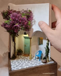 a hand is holding up a miniature house with flowers on the front and side windows