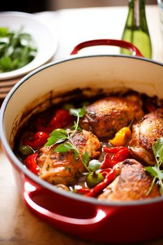 a red pot filled with chicken and vegetables on top of a wooden table next to a bottle of wine