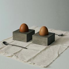 two eggs are sitting on concrete blocks with spoons next to them and a napkin