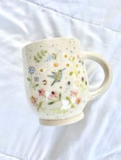 a white coffee cup with flowers painted on the side and stars in the middle, sitting on a bed