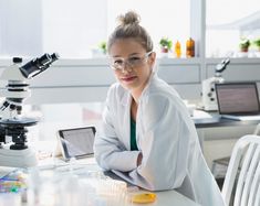 a woman in white lab coat sitting at a table with microscopes and other laboratory equipment