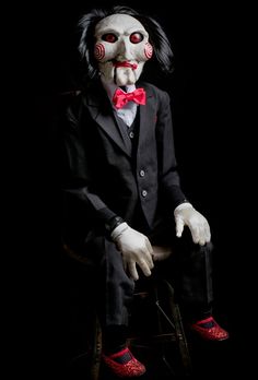 a creepy clown sitting on top of a chair wearing red shoes and a black suit