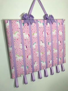 a pink wall hanging with purple ribbons and unicorns on it's sides, attached to a white wall