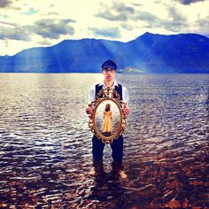 a man standing in the water holding a mirror with mountains in the backgroud