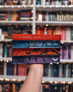 a hand holding three books in front of a book shelf filled with many different colored books