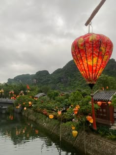 a red lantern is hanging over the water in front of some hills and trees with lights on them