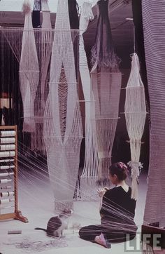 a woman sitting on the ground in front of several hanging objects and a cat looking at her phone