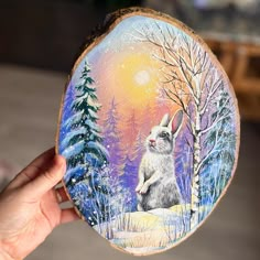 a hand holding up a painting of a rabbit in the snow with trees on it