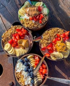 four bowls filled with fruit and granola on top of a wooden table next to spoons