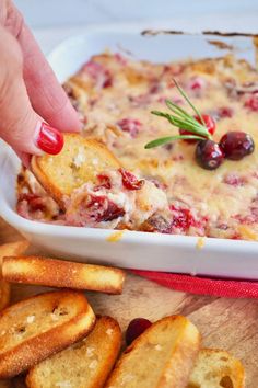 a hand dipping some bread into a casserole with cranberries and olives