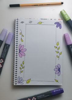 an open notebook surrounded by markers and crayons on a white surface with flowers