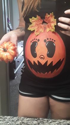 a pregnant woman holding an orange doughnut in front of her belly wearing a black shirt with a pumpkin on it