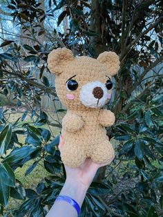 a hand holding a small brown teddy bear in front of some bushes and trees with green leaves