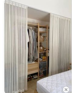 an open closet with white curtains and clothes on shelves next to a bed in a bedroom