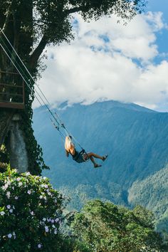 a woman is swinging from a rope in the air with mountains and clouds behind her