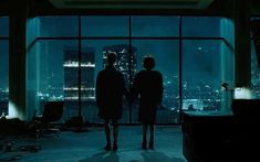 two people standing in front of a window looking out at the city lights and skyscrapers