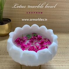 pink flowers are floating in a white bowl on a wooden table next to a potted plant