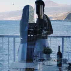 a woman standing on a balcony next to the ocean with a bottle of wine in front of her