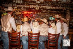 four men in cowboy hats sit at a bar with their backs turned to the camera