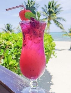 a pink drink sitting on top of a wooden table next to the ocean and palm trees