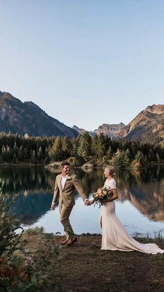 a bride and groom standing next to a lake with mountains in the background at their wedding