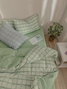 a bed with green sheets and pillows next to a small potted plant on the floor