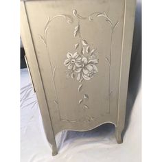 an antique white painted cabinet with flowers on the front and side panels, sitting on a bed sheet