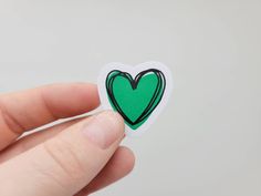 a hand holding a green heart sticker in front of a white background with black outline