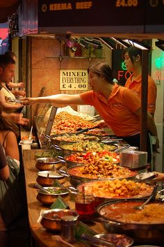 people are standing at a buffet table with many different foods on it and one person is reaching for something