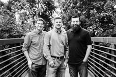 three men are standing on a bridge posing for a black and white photo with trees in the background