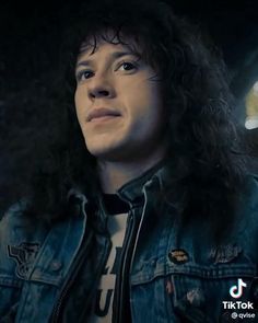 a man with long hair wearing a denim jacket and looking off to the side in a dark room