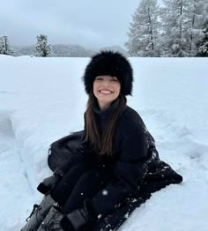 a woman sitting in the snow wearing a black coat and hat with fur on it