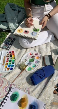 a woman is sitting on the grass painting with watercolors and other art supplies