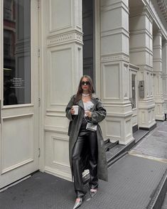 a woman standing on the sidewalk in front of a building wearing black leather pants and a gray trench coat