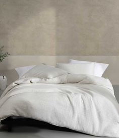 an unmade bed with white linens and pillows on the headboard, in front of a concrete wall