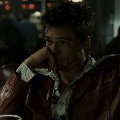 a man in a leather jacket sitting at a table with a drink and cell phone