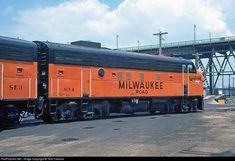 an orange and black train sitting on the tracks next to a large bridge over water