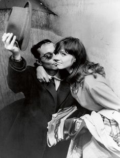 black and white photograph of man kissing woman's face with hat on head, in front of wall