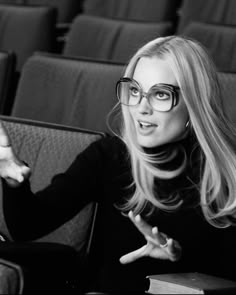 a woman with glasses sitting in front of an empty theater seat and pointing to the side