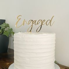 a white cake with gold lettering on top and a plant in the corner behind it